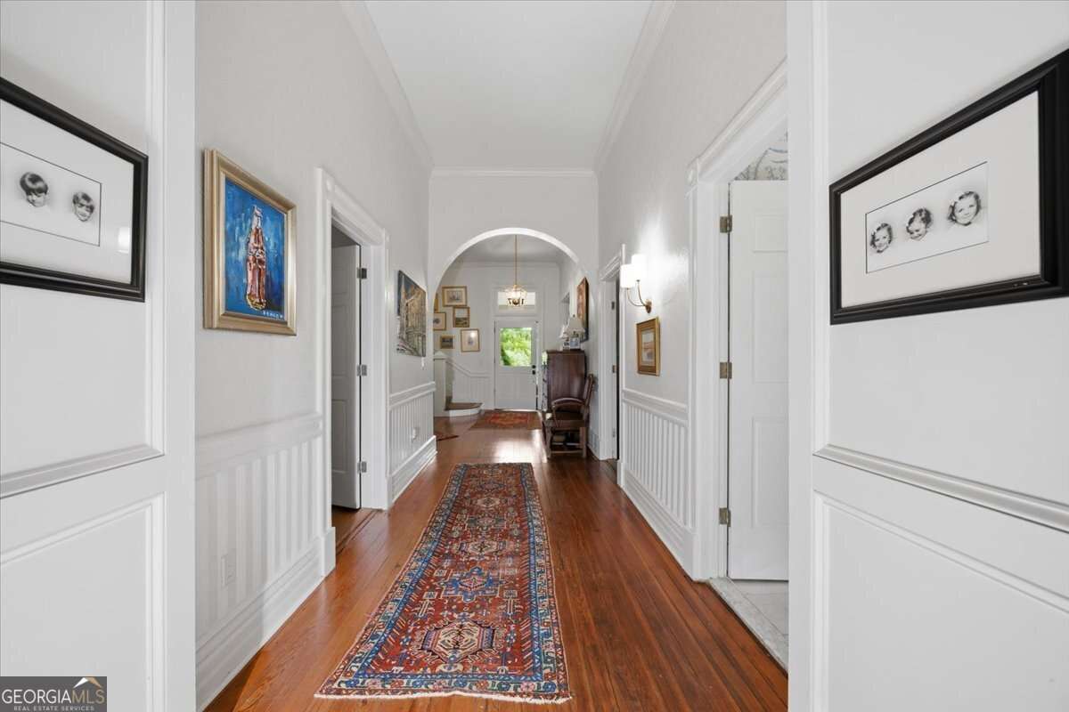 Long, bright hallway with hardwood floors, white wainscoting, high ceilings, and decorative artwork, leading to an arched doorway and entry area.