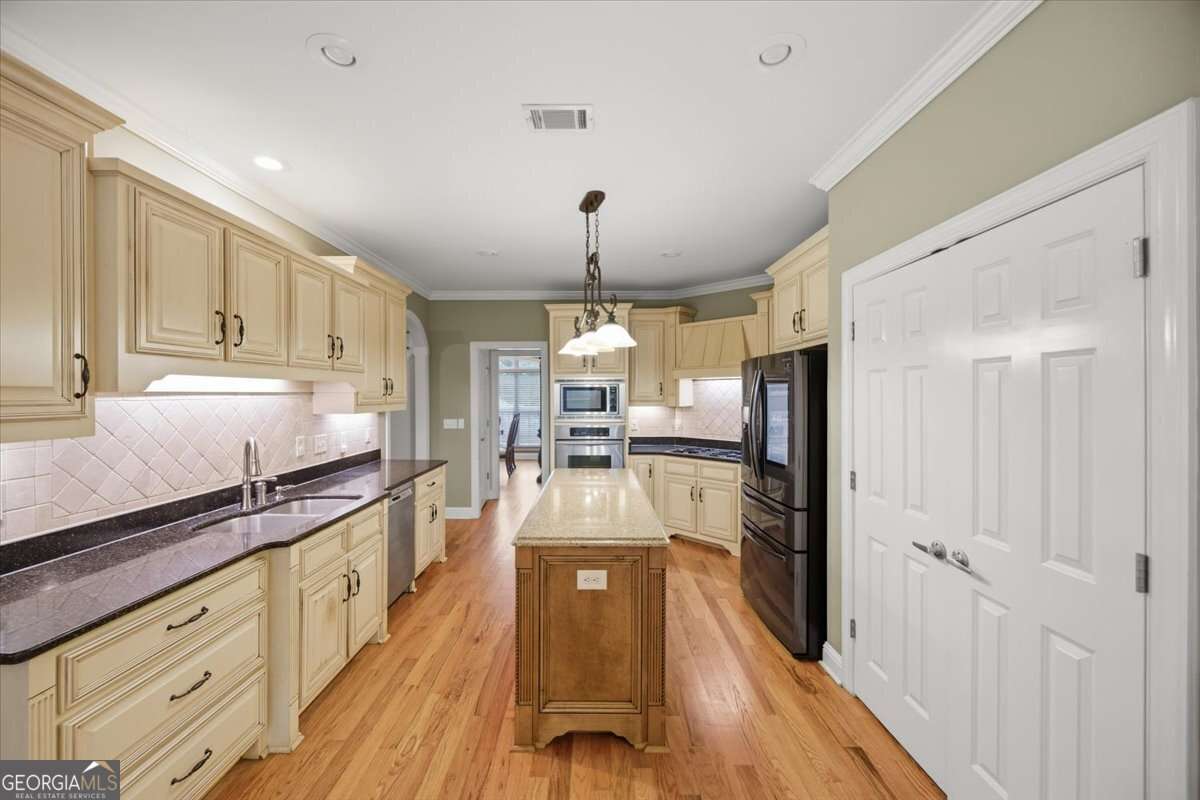 Long kitchen with cream cabinets, granite counters, and a central island; stainless steel appliances and hardwood floors describe a bright, modern cooking space.