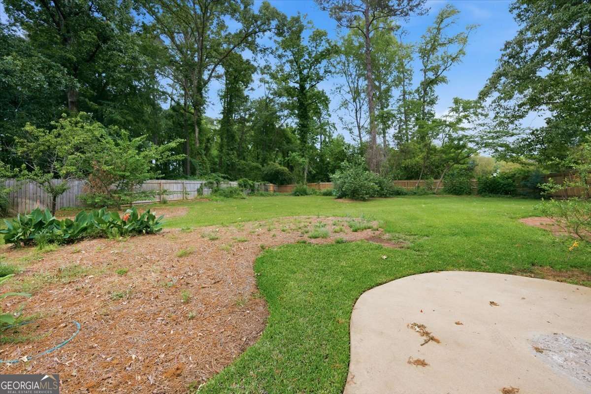 Backyard with green lawn, mulched garden bed on left, and tall trees behind a wooden fence.