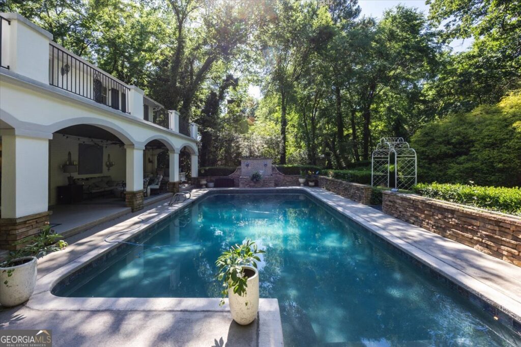 Resort-style pool at 4717 Brae Burn Lane in Macon GA surrounded by mature trees, stone privacy walls, and covered outdoor living space beneath an elegant terrace balcony.