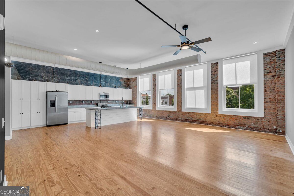 Renovated open-concept living and kitchen area at 565 Poplar Street in Macon, GA, featuring hardwood floors, exposed brick walls, high ceilings, large windows, and stainless steel appliances.