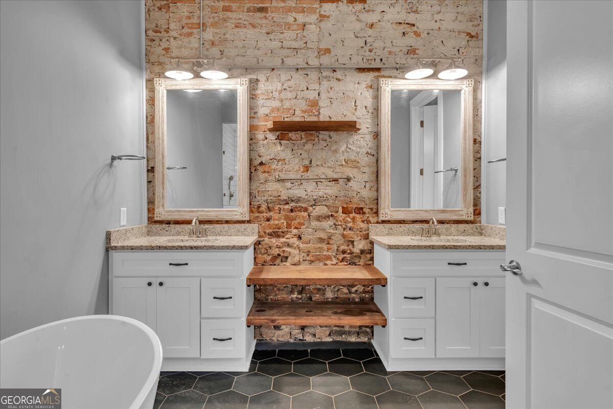 Renovated bathroom at 565 Poplar Street in Macon, GA, featuring dual vanities, granite countertops, exposed historic brick, hexagon tile flooring, and modern finishes.